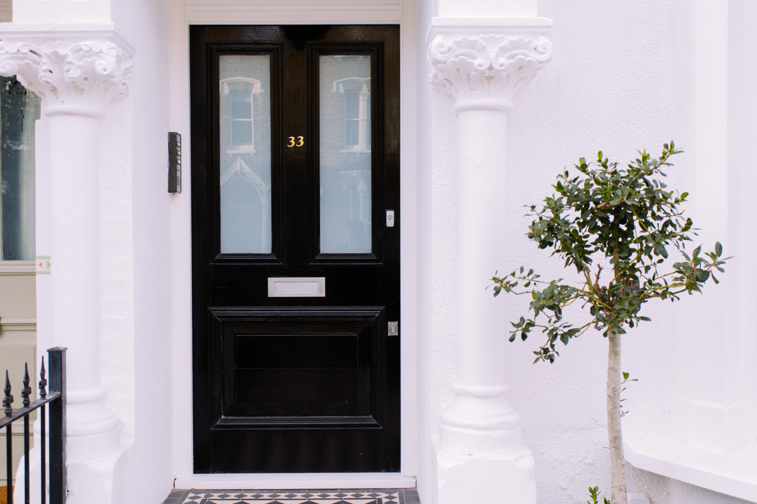 A picture of the front of a property with a gloss black front door, two white pillars either side of the porch and a small tree. 